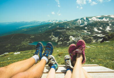 Three people relax on a grassy hilltop, their legs stretched out and feet in sneakers, overlooking a scenic mountain landscape with lakes and patches of snow under a clear blue sky.