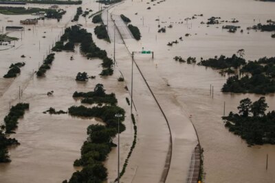 Aerial view of a major highway and surrounding land submerged in floodwater, with only tree tops and road signs visible above the water.