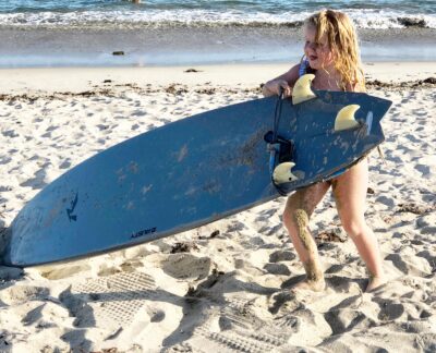 A young child with wet, sandy hair struggles to carry a large surfboard on a sandy beach, with the ocean and waves visible in the background.