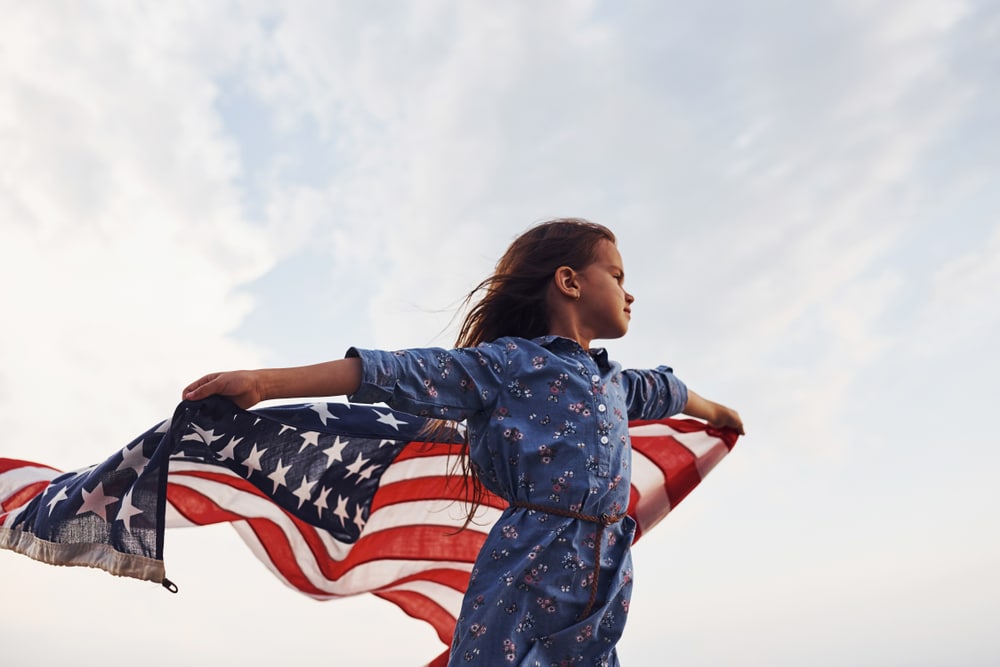 Patriotic,Female,Kid,With,American,Flag,In,Hands.,Against,Cloudy