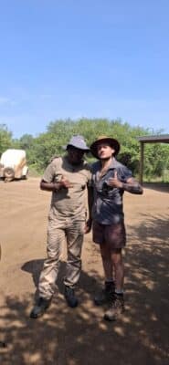 Two men stand side by side outdoors on a dirt area, both giving thumbs-up gestures. They are wearing hats and casual outdoor clothing. Green trees and clear blue sky are visible in the background.