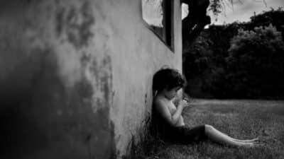 A young child sits barefoot on the grass, leaning against an old, weathered wall outside, intently examining something in their hands. The scene is in black and white and feels quiet and contemplative.
