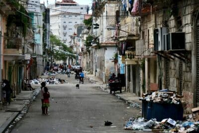 A rundown urban street with garbage scattered along the sidewalks and overflowing bins. People, including children, walk and stand in the street. Buildings on both sides appear old and worn.