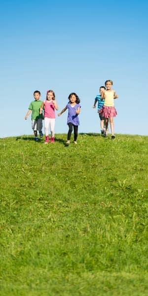 Five children are running down a grassy hill under a clear blue sky, wearing colorful clothes and smiling, enjoying a sunny day outdoors.