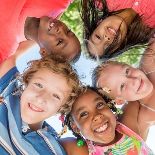 Five children of diverse backgrounds stand in a circle outdoors, smiling and looking down at the camera. Their heads are close together, with arms around each other, and green foliage is visible in the background.