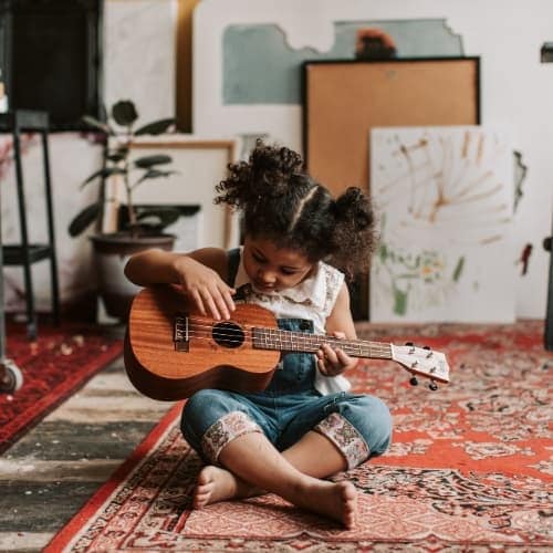 A young girl sitting cross-legged on a patterned rug indoors, focused on playing a small wooden guitar or ukulele, surrounded by art supplies and canvases in a cozy creative space.