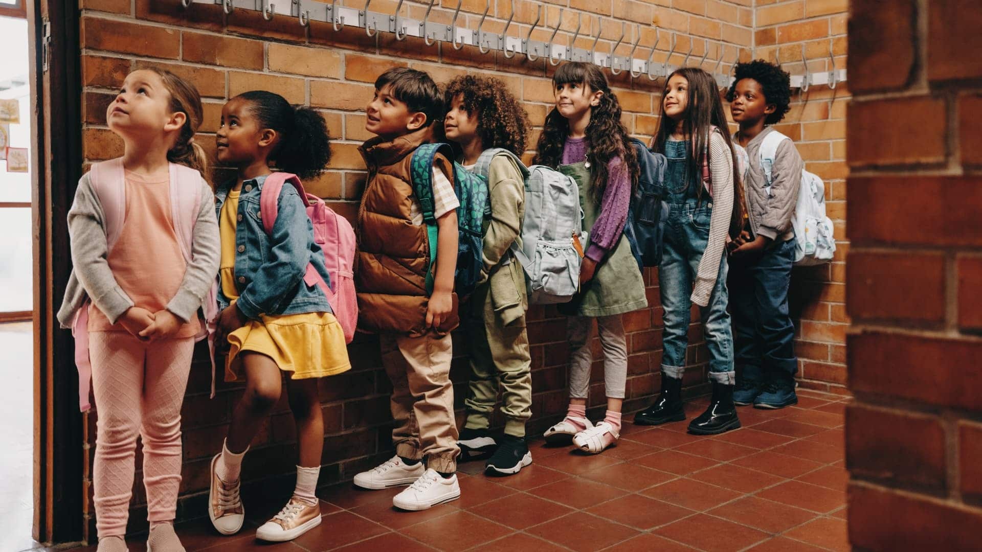Seven young children with backpacks stand in a line against a brick wall in a school hallway, facing left and appearing to wait patiently.