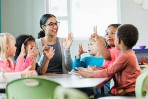 A teacher sits at a table with a group of young children, all raising their hands and smiling, engaging in an interactive classroom activity.