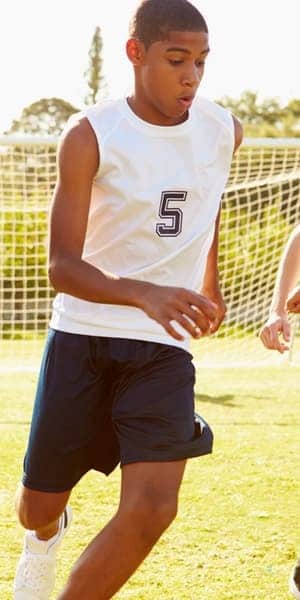 Teen boy in a white sleeveless shirt with the number 5 and navy shorts plays soccer on a grassy field, with a goal net and trees visible in the background.