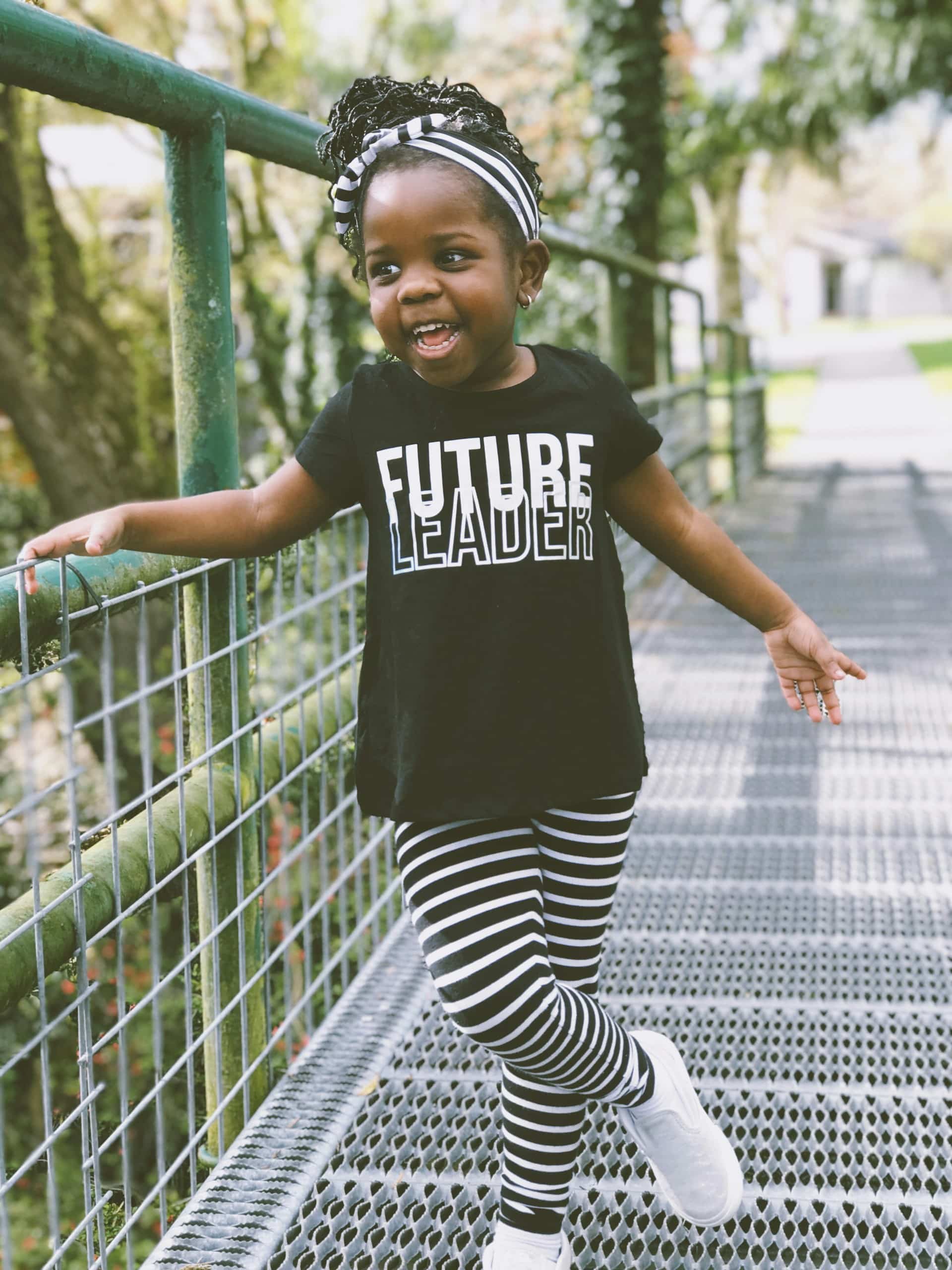 Smiling young girl in striped leggings and a Future Leader shirt stands on a metal walkway, holding the railing, with trees and a house in the background.