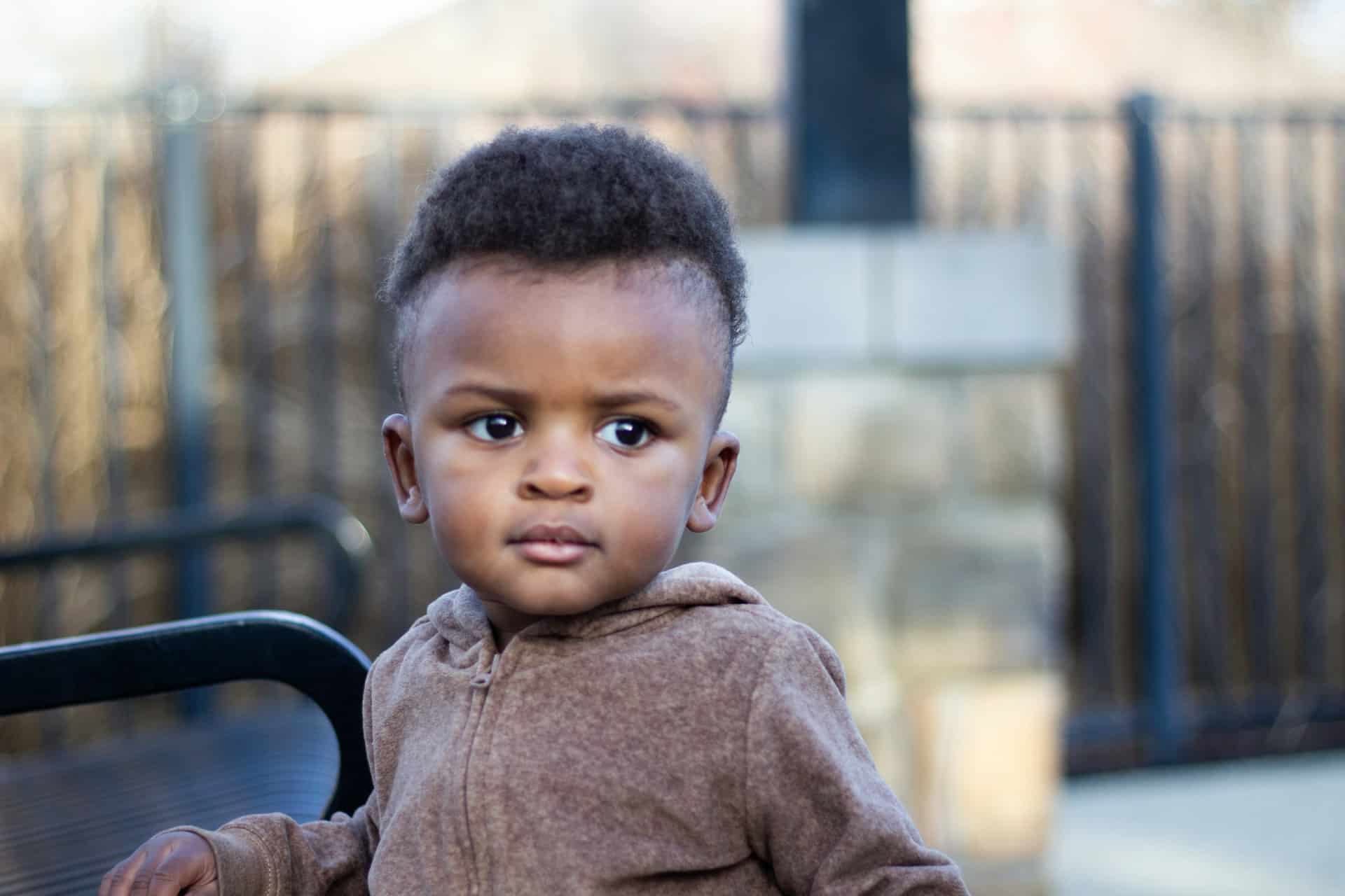 A young child with short curly hair wearing a brown hoodie looks slightly to the side with a serious expression, sitting outdoors near a black metal bench and a stone fence.
